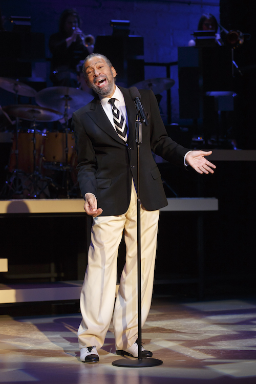 Maurice Hines sings into a microphone with a live band behind him